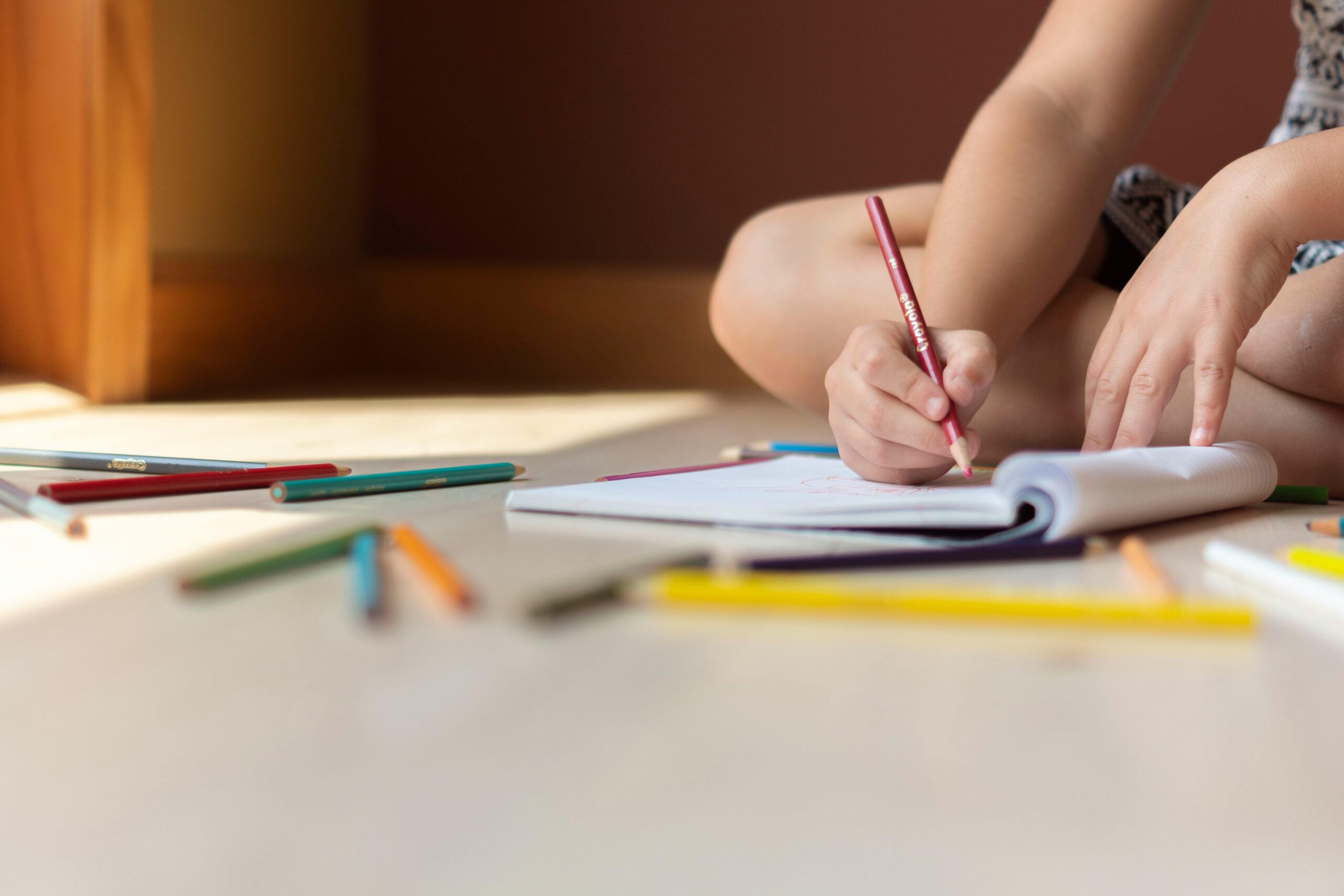 Crop kid sitting on floor and writing in notebook